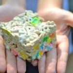 Child's hands holding a colorful Marshmallow Krispy Treats Used for Bribery square with rainbow sprinkles and cereal coating