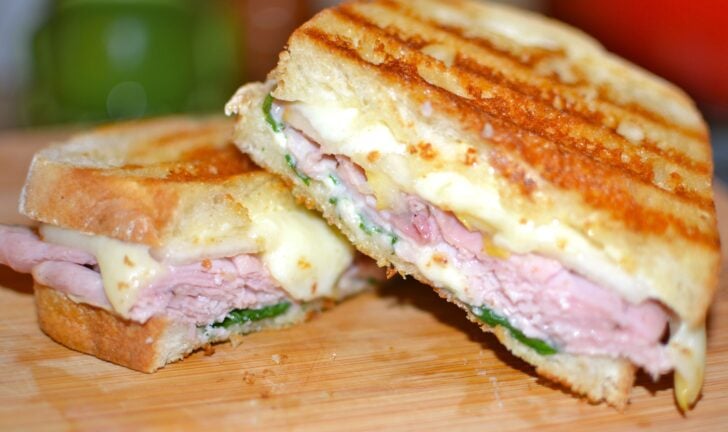 Ham & Cheese Panini with Pears & Spinach cut in half on wooden board, showing melted cheese, sliced ham, and green spinach between toasted b