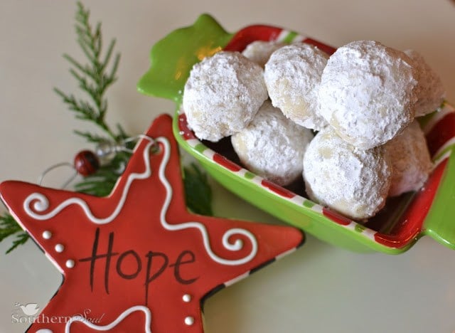 Powdered sugar-coated Mexican Wedding Cookies in a festive red and green container with holiday decorations and rosemary sprig