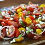 Heirloom Tomato Salad with sliced red tomatoes, yellow peppers, red onions, and crumbled feta cheese on a wooden plate