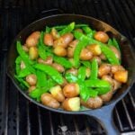 Grill Roasted Peas & Potatoes cooking in cast iron skillet on grates, showing browned potatoes and bright green pea pods