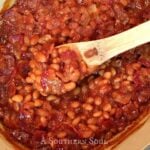 Southern Baked Beans simmering in a Dutch oven with a wooden spoon stirring the red sauce and beans together
