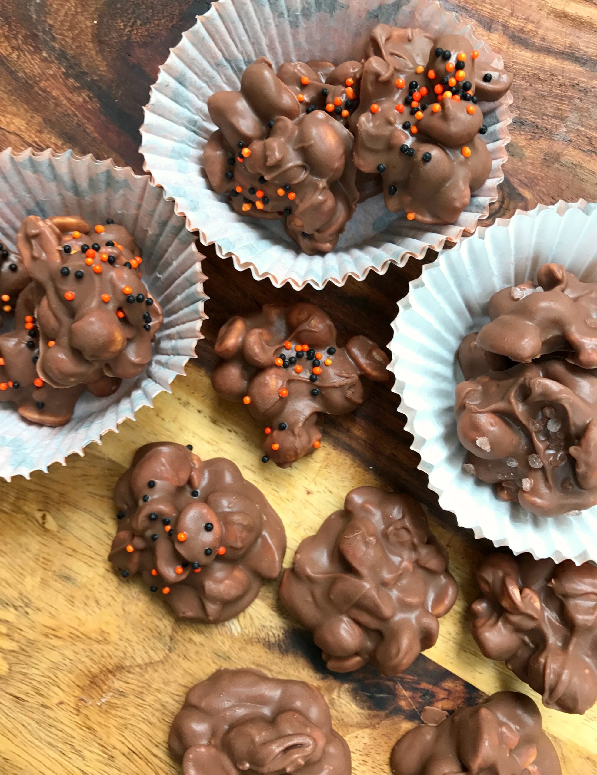 Chocolate Peanut Crock Pot Candy clusters on wood surface in white cupcake liners, topped with orange and black sprinkles