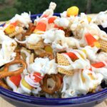 Halloween Crunch snack mix in blue bowl with pretzels, cereal, popcorn, candy corn and orange and yellow candies outdoors