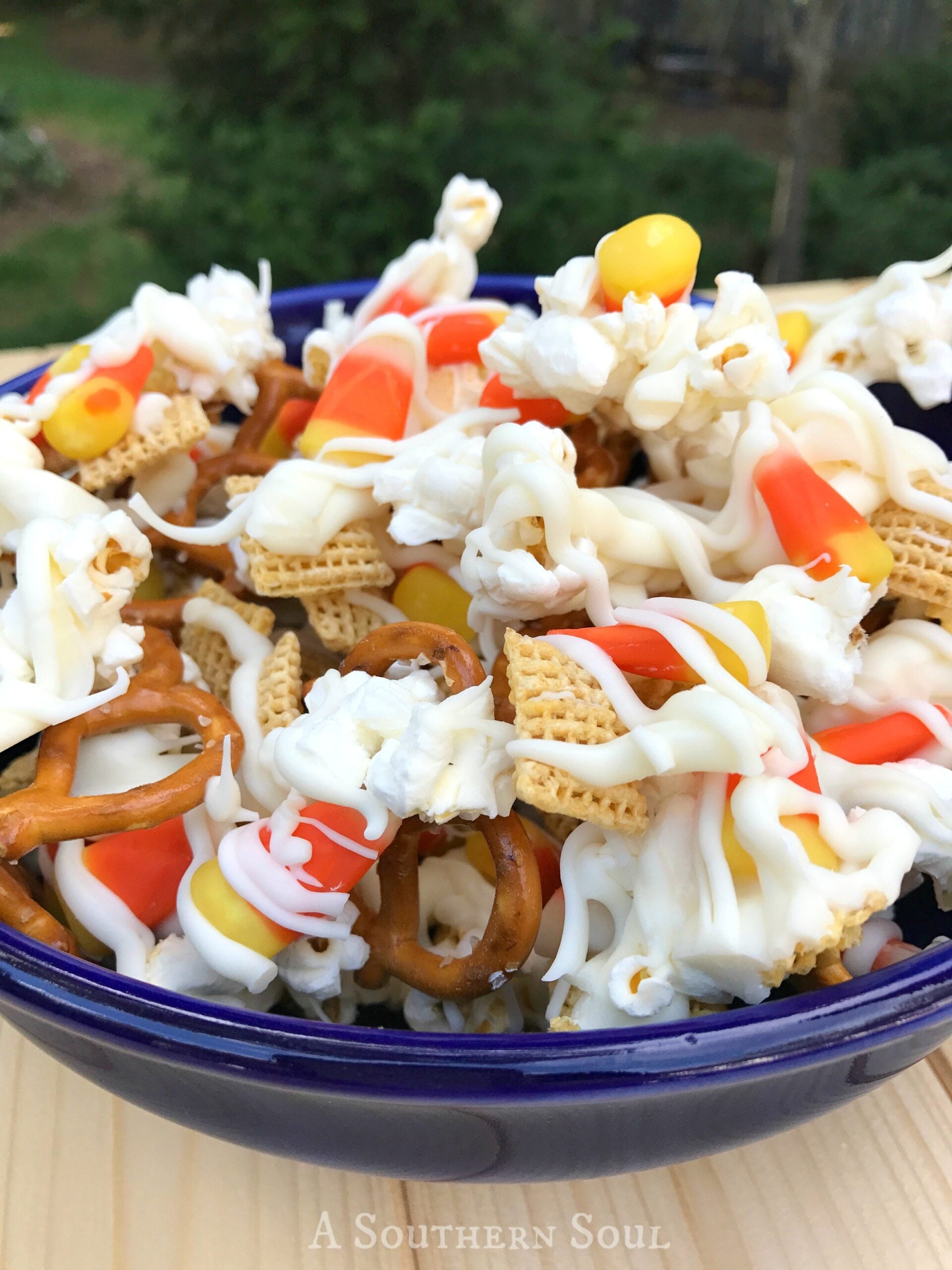 Halloween Crunch snack mix in blue bowl with pretzels, cereal, popcorn, candy corn and orange and yellow candies outdoors