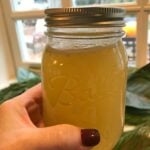 Hand holding jar of golden Crock Pot Chicken Stock | Liquid Gold with metal lid, windows and greenery visible in background