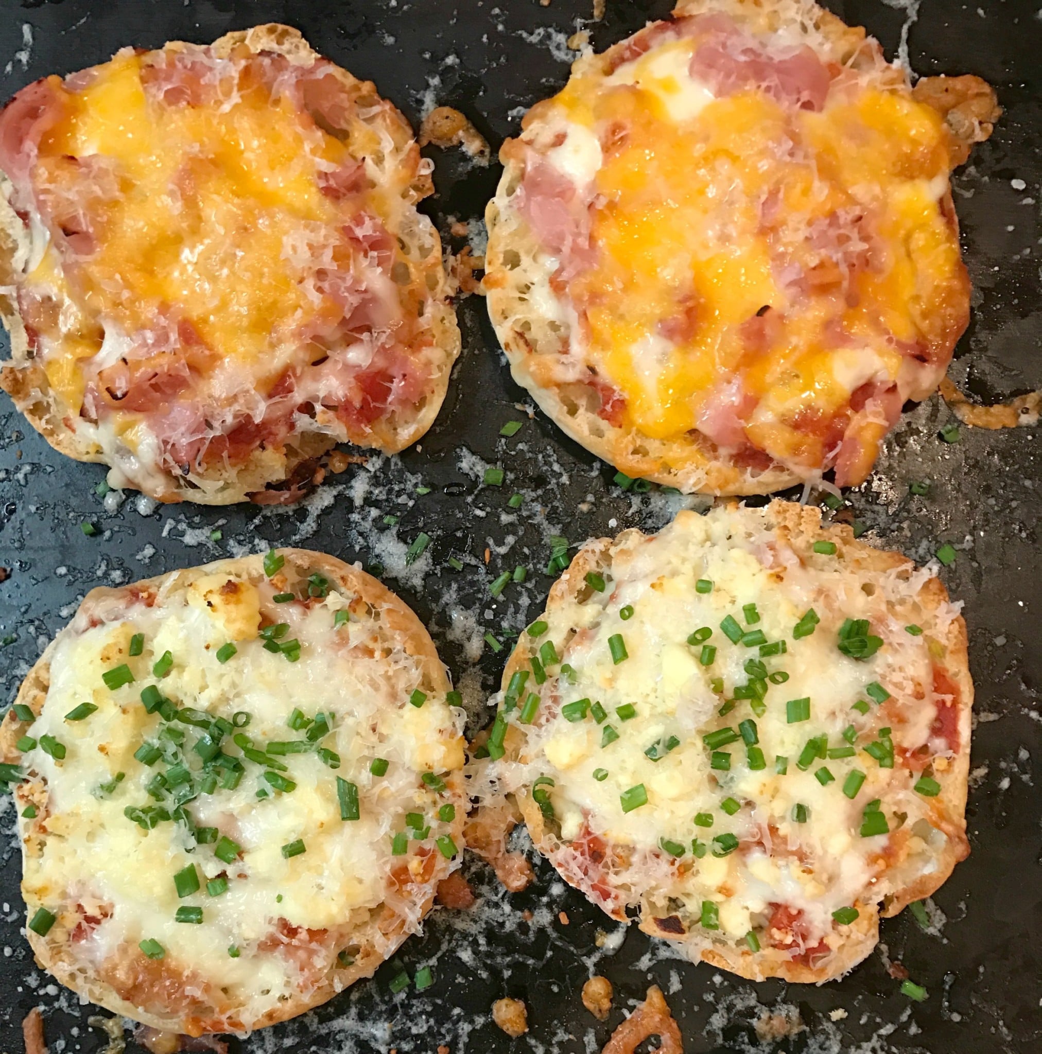 Four English Muffin Pizzas on dark griddle topped with melted cheese, ham, and green onions, two varieties shown