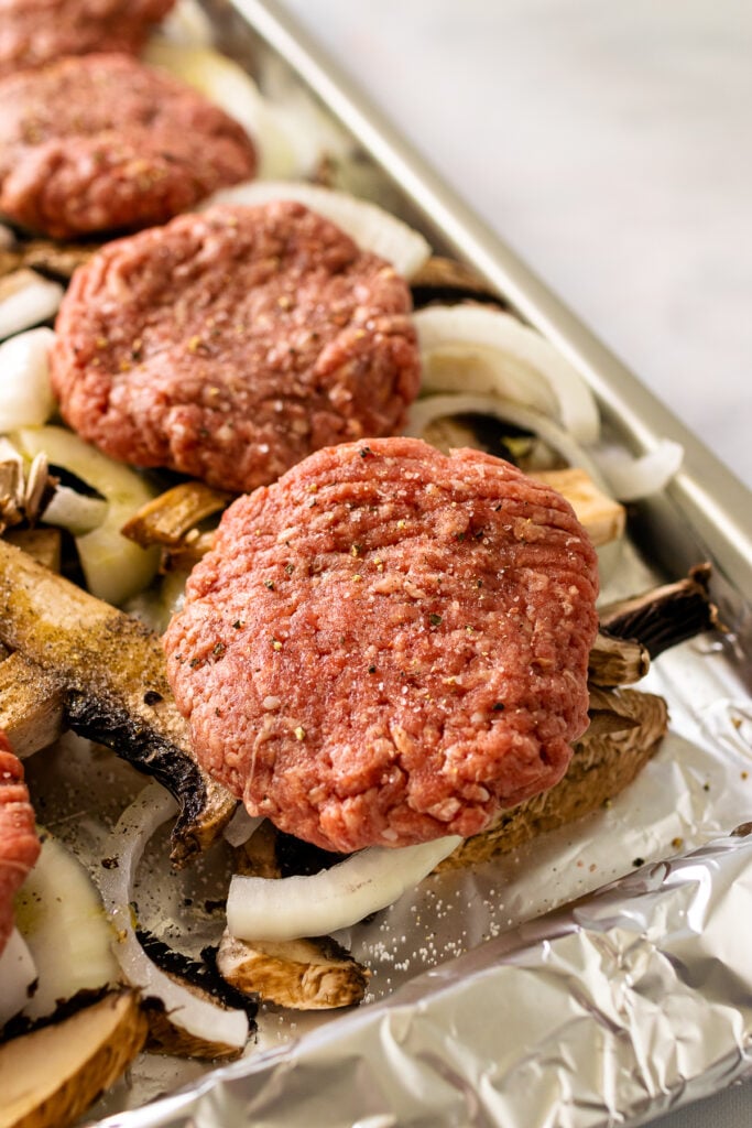 Uncooked hamburger steak patties placed on a sheet pan over a layer of raw mushrooms and onions, ready to be cooked.