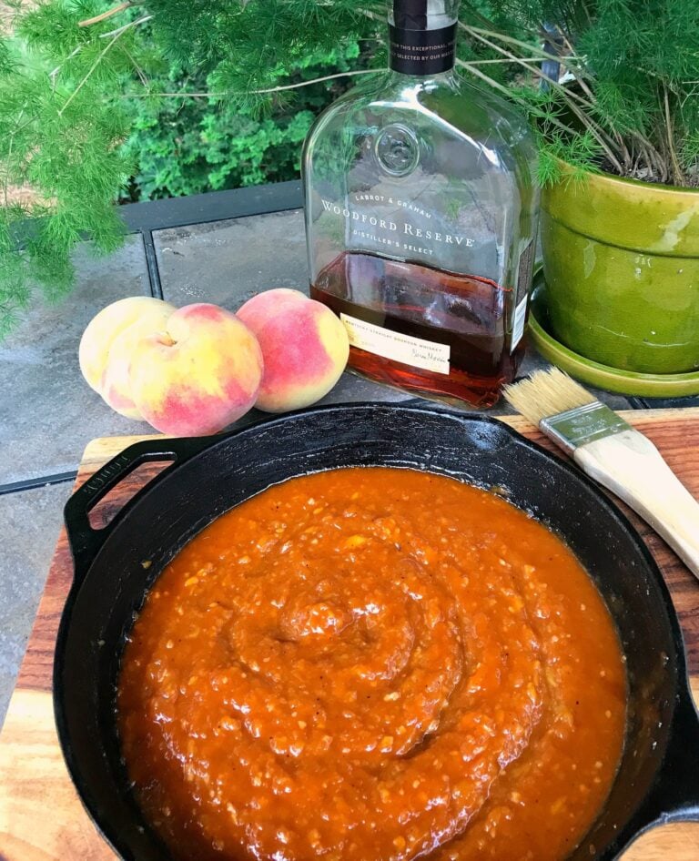 Peachy Bourbon BBQ Sauce simmering in cast iron skillet with fresh peaches and Woodford Reserve bourbon bottle displayed outdoors