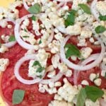 Tomato and corn summer salad with fresh mozzarella, red onion slices, basil, and black pepper on yellow background