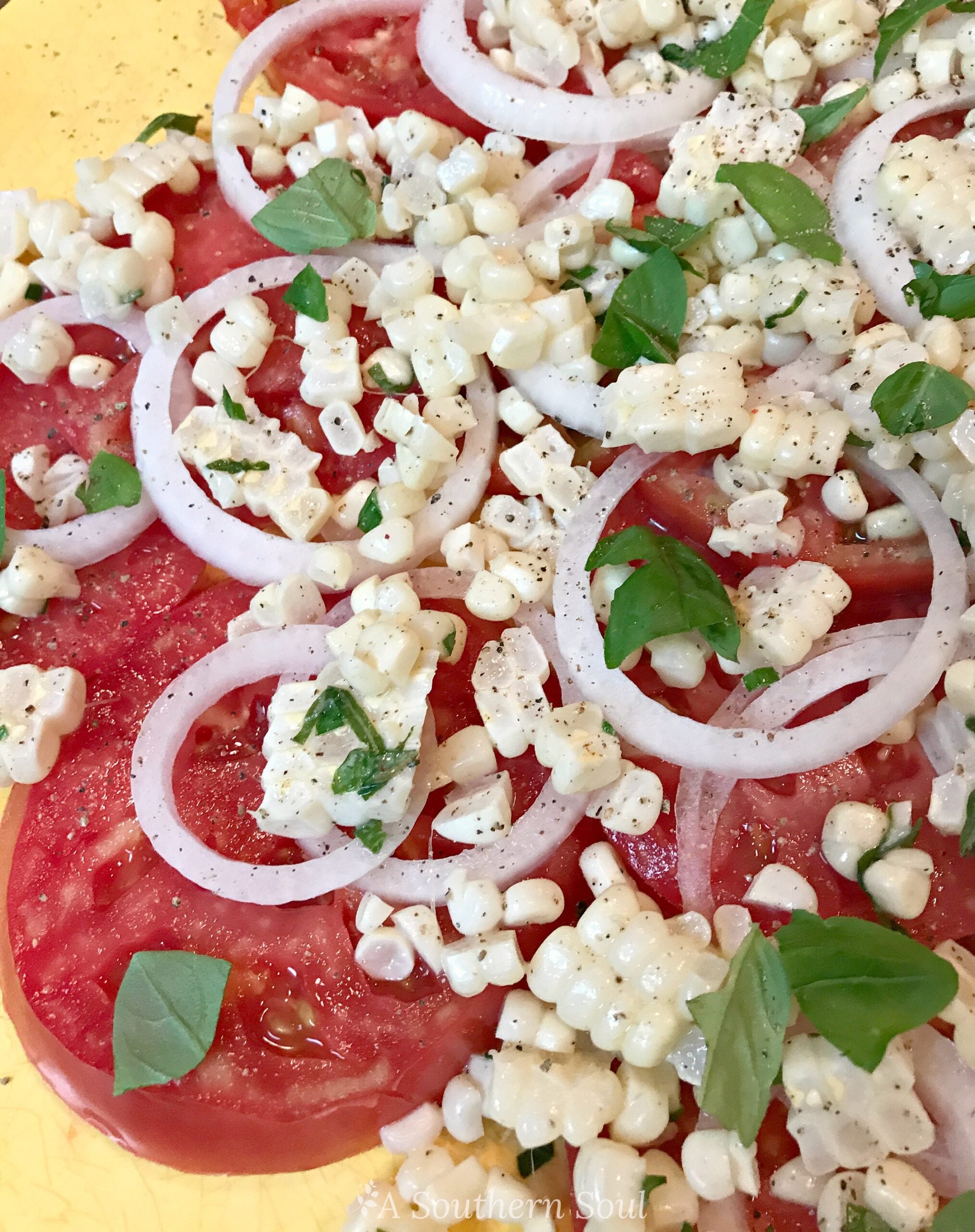 Tomato and corn summer salad with fresh mozzarella, red onion slices, basil, and black pepper on yellow background