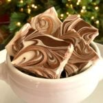Tiger Butter Fudge pieces with chocolate and vanilla swirls stacked in a white ceramic bowl against a blurred Christmas tree background
