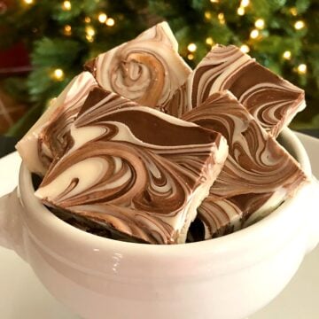Tiger Butter Fudge pieces with chocolate and vanilla swirls stacked in a white ceramic bowl against a blurred Christmas tree background