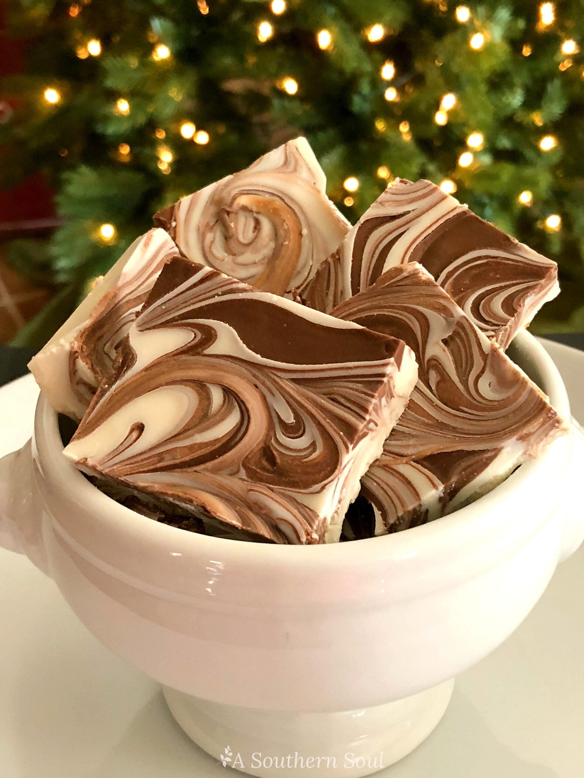 Tiger Butter Fudge pieces with chocolate and vanilla swirls stacked in a white ceramic bowl against a blurred Christmas tree background
