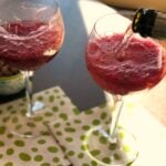 Two Blood Orange Mimosas in wine glasses with ice on a green polka dot napkin, one being poured with a bottle