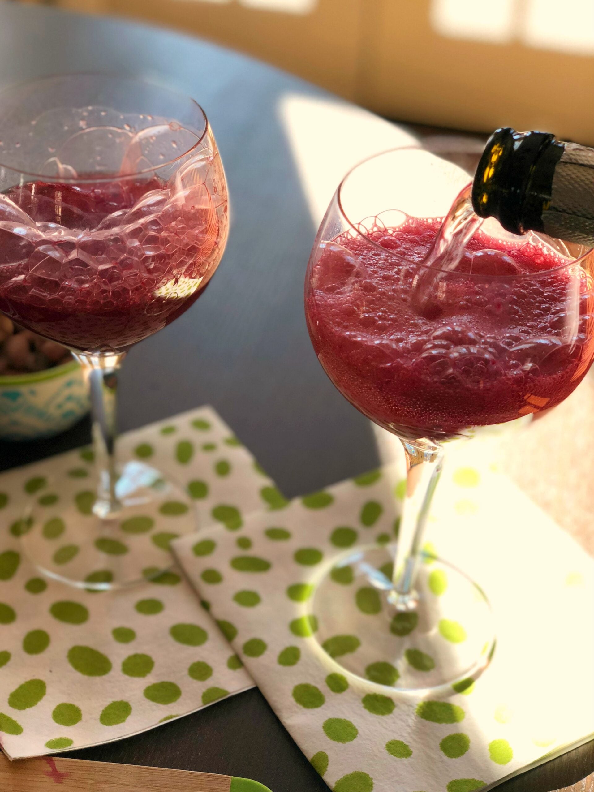 Two Blood Orange Mimosas in wine glasses with ice on a green polka dot napkin, one being poured with a bottle