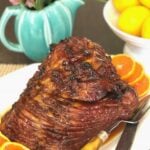 Citrus Molasses Glazed Ham with glossy brown coating, orange slices, lemons, and pink flowers on white plate with fork