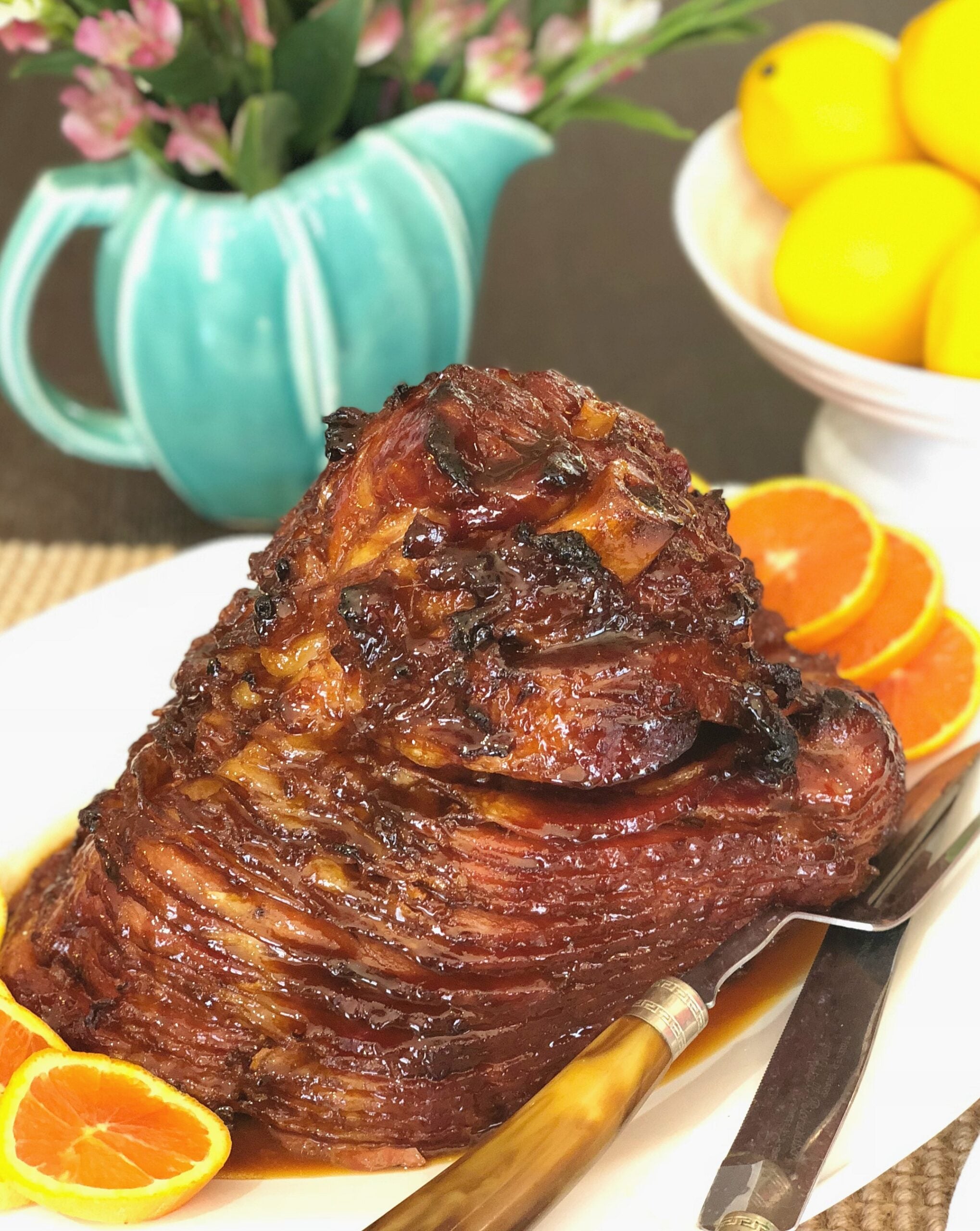 Citrus Molasses Glazed Ham with glossy brown coating, orange slices, lemons, and pink flowers on white plate with fork