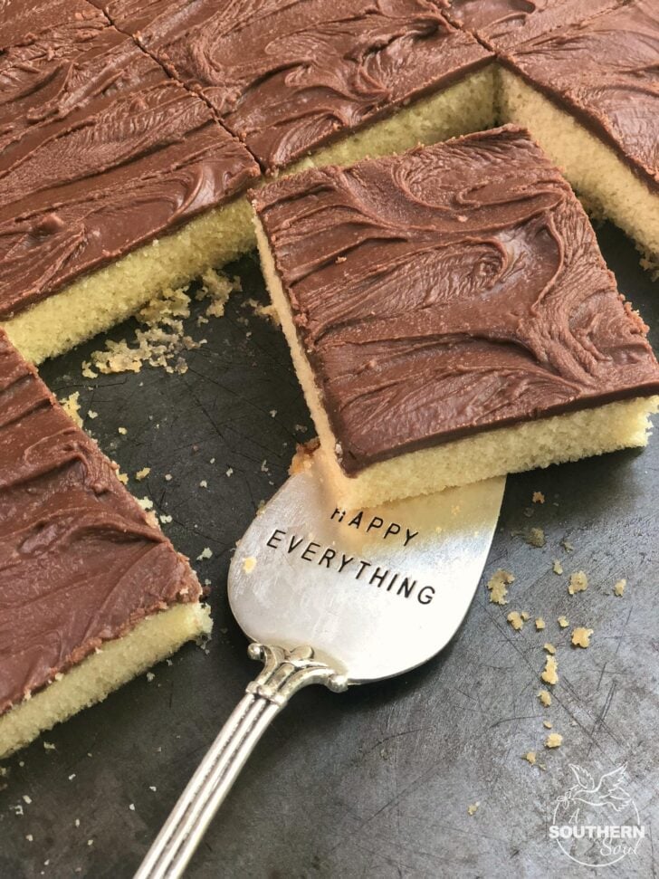 Vanilla Sheet Cake with Chocolate Fudge Icing sliced into squares with a silver spoon reading Happy Everything on dark surface