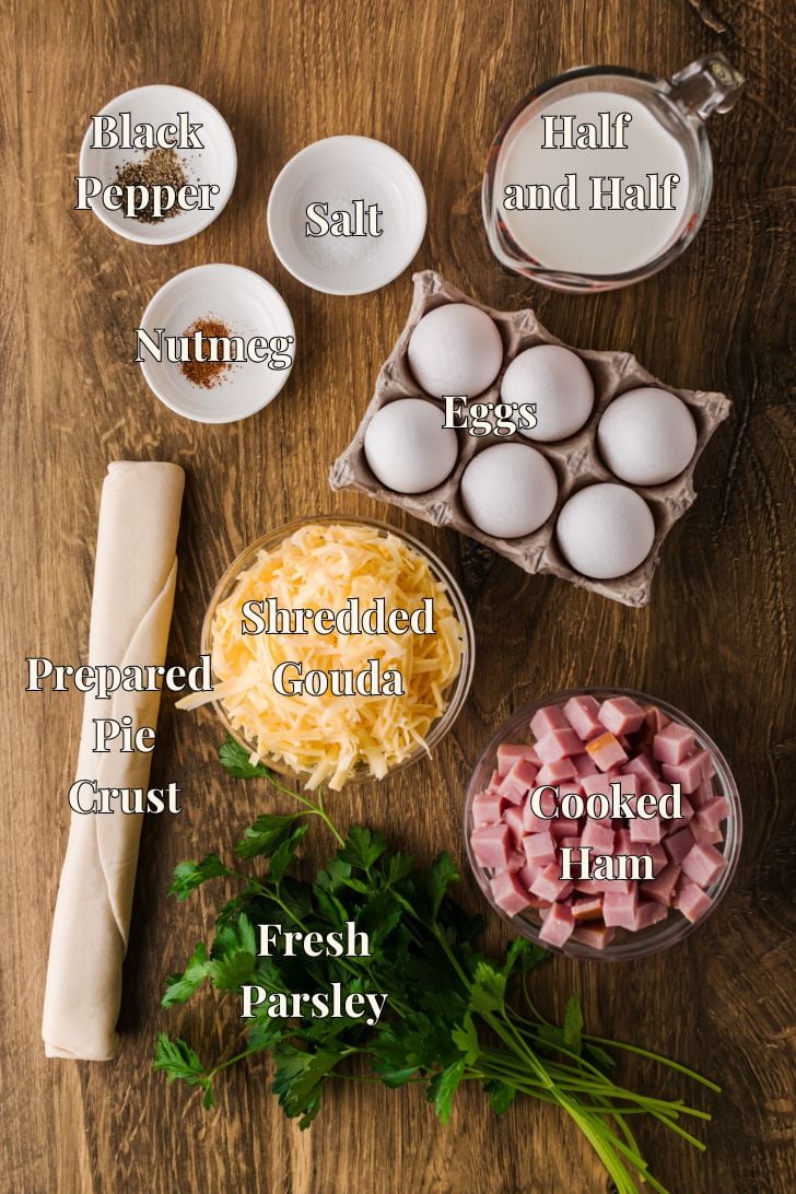 Ingredients for ham and cheese quiche arranged on the counter, including eggs, half-and-half, diced ham, shredded gouda, and a deep-dish pie crust.