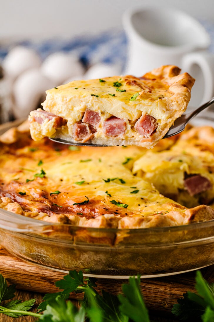 A slice of ham and cheese quiche lifted from the pie dish on a serving spatula, showing the creamy filling and melty cheese inside.