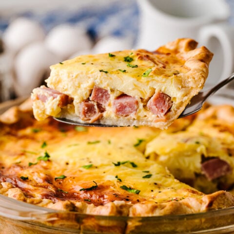 Ham and Cheese Quiche slice on server in glass pie dish, garnished with chives, eggs and pitcher blurred in background