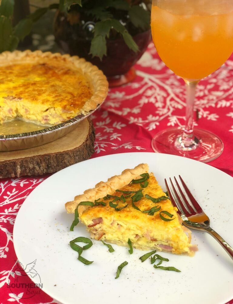Slice of Ham and Cheese Quiche on white plate garnished with green onions, fork beside, whole quiche and orange juice in background
