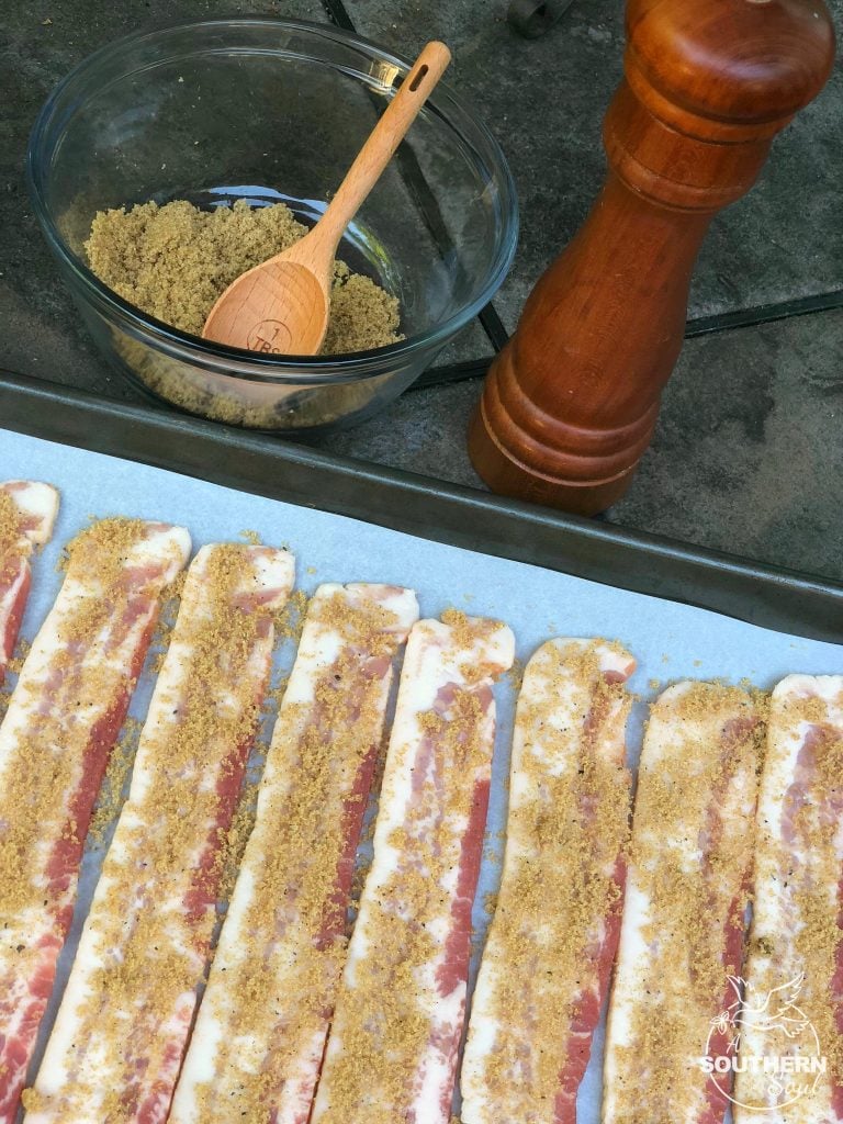 Glass bowl wit brown sugar, a pepper mill and slices of bacon with brown sugar on a parchment lined baking sheet.