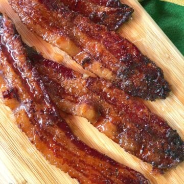 Brown Sugar Pepper Candied Bacon strips on wooden board with green napkin, glistening with caramelized coating and charred edges