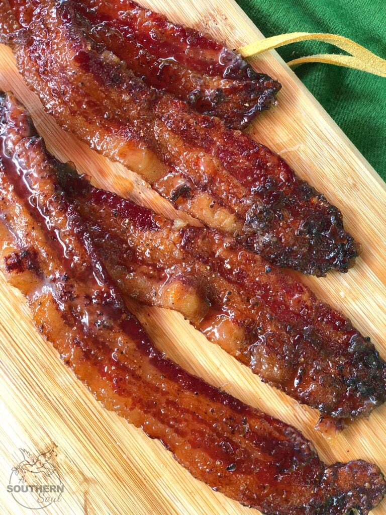 Brown Sugar Pepper Candied Bacon strips on wooden board with green napkin, glistening with caramelized coating and charred edges