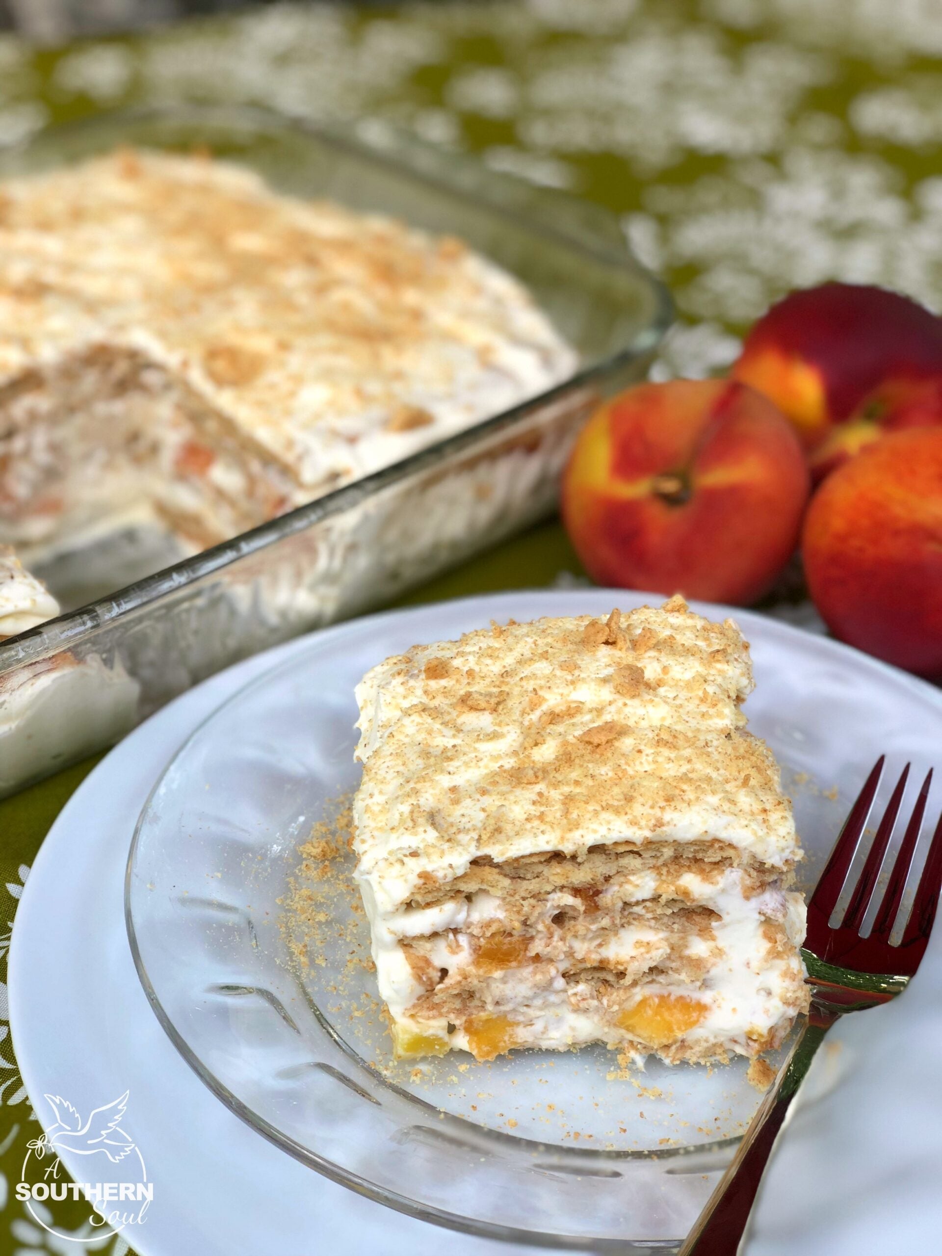 Slice of Peaches & Cream Icebox Cake on white plate with fork, showing layers of wafers, peach filling, and cream frosting topped with crumb