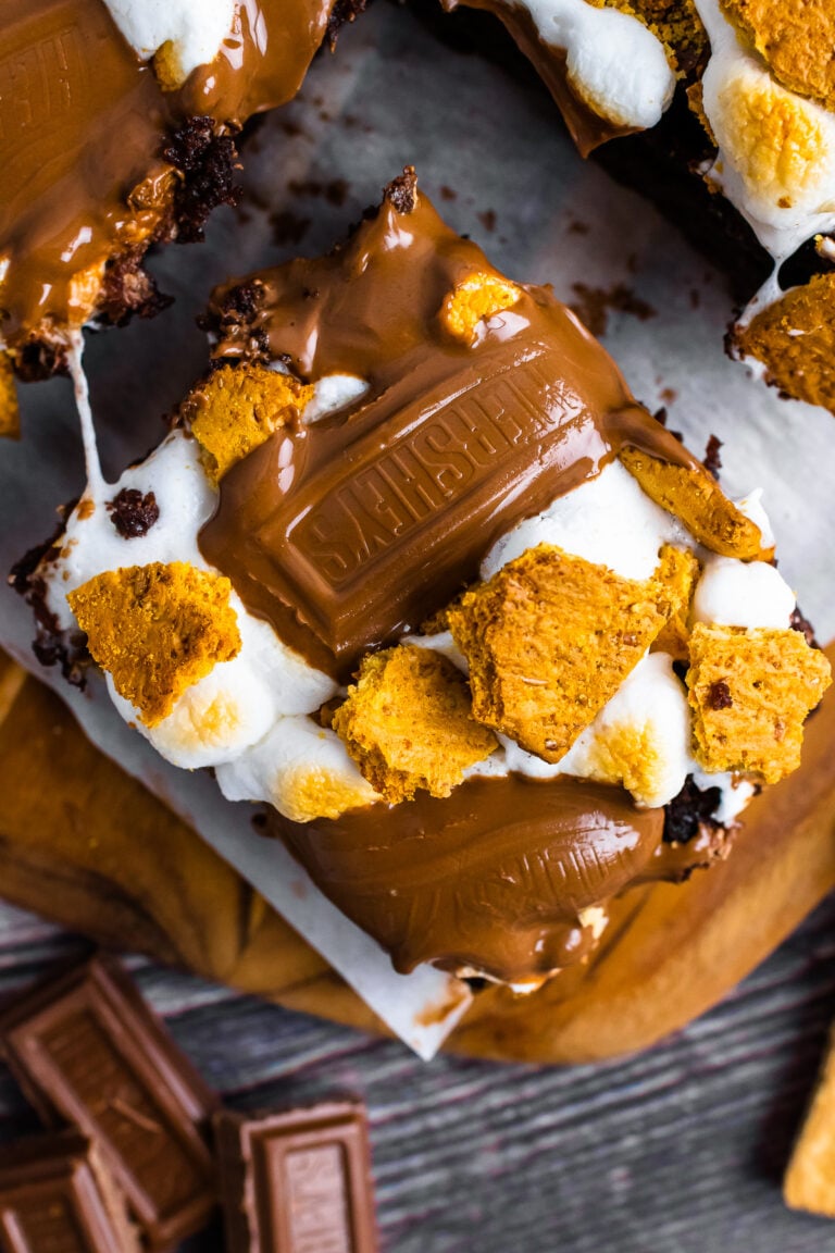 Super Simple S'mores Brownies stacked on parchment with melted chocolate, toasted marshmallows, and golden graham cracker pieces on dark woo