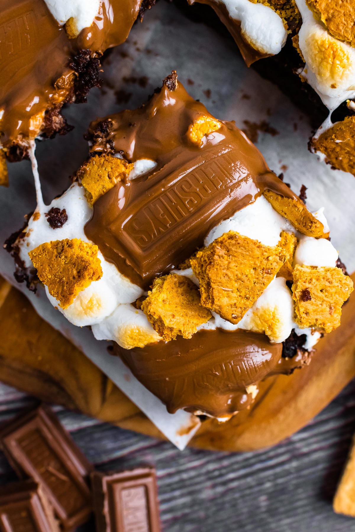 Super Simple S'mores Brownies stacked on parchment with melted chocolate, toasted marshmallows, and golden graham cracker pieces on dark woo