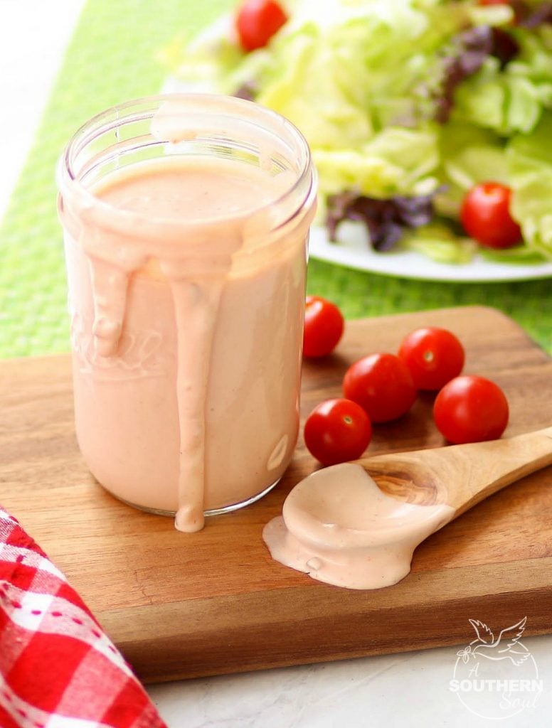 A wooden spoon has been dipped into this rich, flavorful, creamy Thousand Island Dressing, and then set to the side as the dressing spills over the sides of the mason jar it's in.