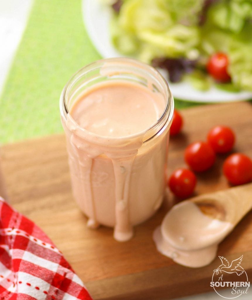 A wooden spoon has been dipped into this rich, flavorful, creamy Thousand Island Dressing, and then set to the side as the dressing spills over the sides of the mason jar it's in.