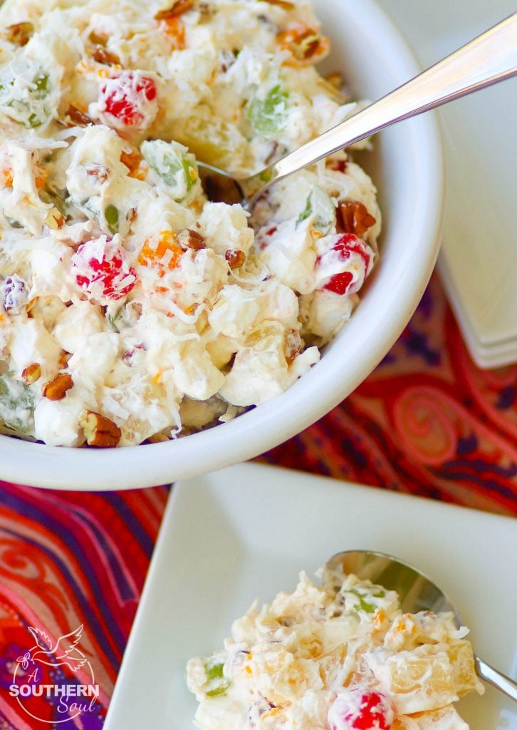 A full, large white serving bowl sits next to a small square plate piled high with Ambrosia Salad, made with oranges, cherries, pineapple and marshmallows is tossed in a light fluffy mixture of fresh whipped cream with a touch of sour cream for extra flavor.