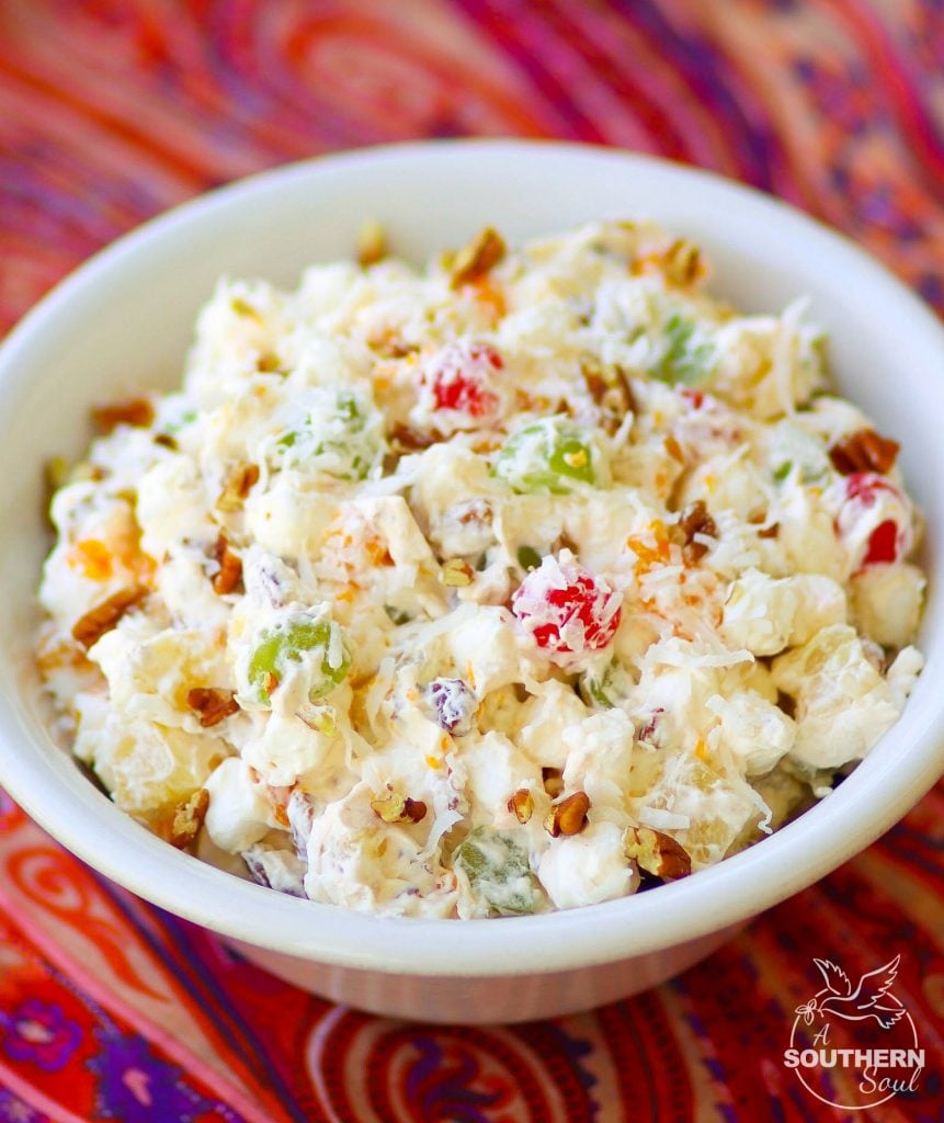 Inside a white serving bowl sits a delicious Ambrosia Salad, made with oranges, cherries, pineapple and marshmallows is tossed in a light fluffy mixture of fresh whipped cream with a touch of sour cream for extra flavor.