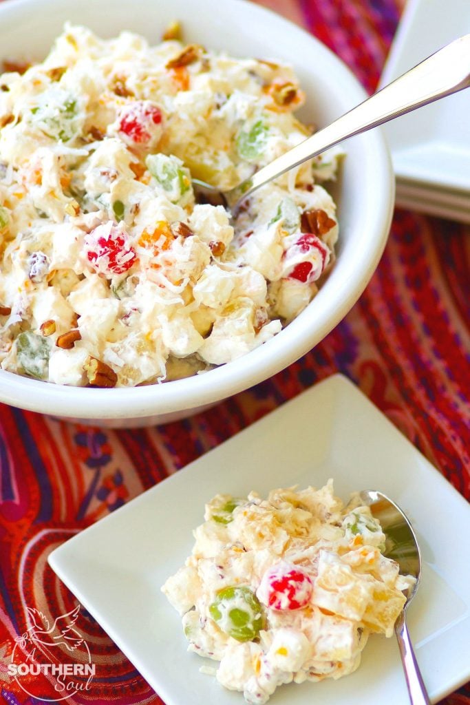 A full, large white serving bowl sits next to a small square plate piled high with Ambrosia Salad, made with oranges, cherries, pineapple and marshmallows is tossed in a light fluffy mixture of fresh whipped cream with a touch of sour cream for extra flavor.