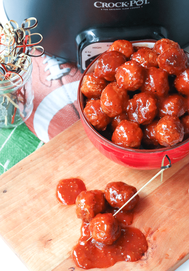 A heaping bowl of hearty meatballs, cooked low and slow in the Crock Pot in a sauce made with BBQ sauce, apricot preserves and whole grain mustard. In front of the bowl sits three individual meatballs with a cocktail toothpick in one of them, and the Crock Pot sides behind the bowl.
