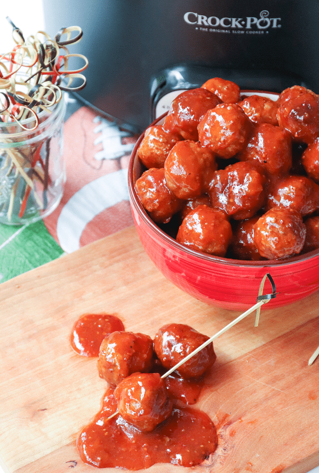A heaping bowl of hearty meatballs, cooked low and slow in the Crock Pot in a sauce made with BBQ sauce, apricot preserves and whole grain mustard. In front of the bowl sits two individual meatballs with cocktail toothpicks in them, and the Crock Pot sides behind the bowl.