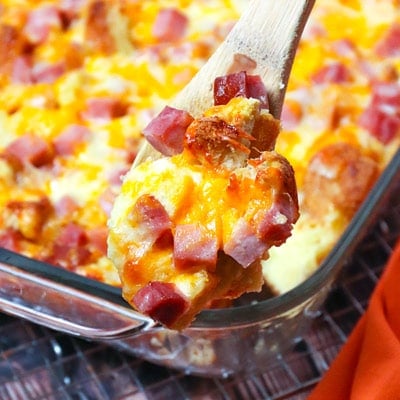 Spatula lifting a golden ham and cheese strata from a baking dish, showing melted cheese and diced ham pieces