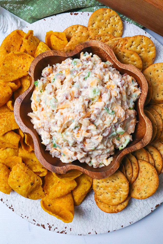 Million Dollar Dip in wooden bowl surrounded by cheese crackers and chips on white plate, overhead view
