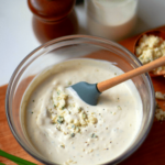 Easy Blue Cheese Dressing in glass bowl with crumbled cheese and black pepper, wooden spoon stirring, ingredients visible behind