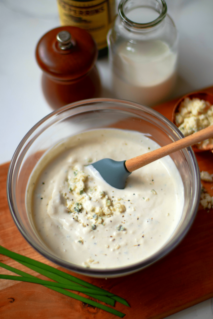 Easy Blue Cheese Dressing in glass bowl with crumbled cheese and black pepper, wooden spoon stirring, ingredients visible behind