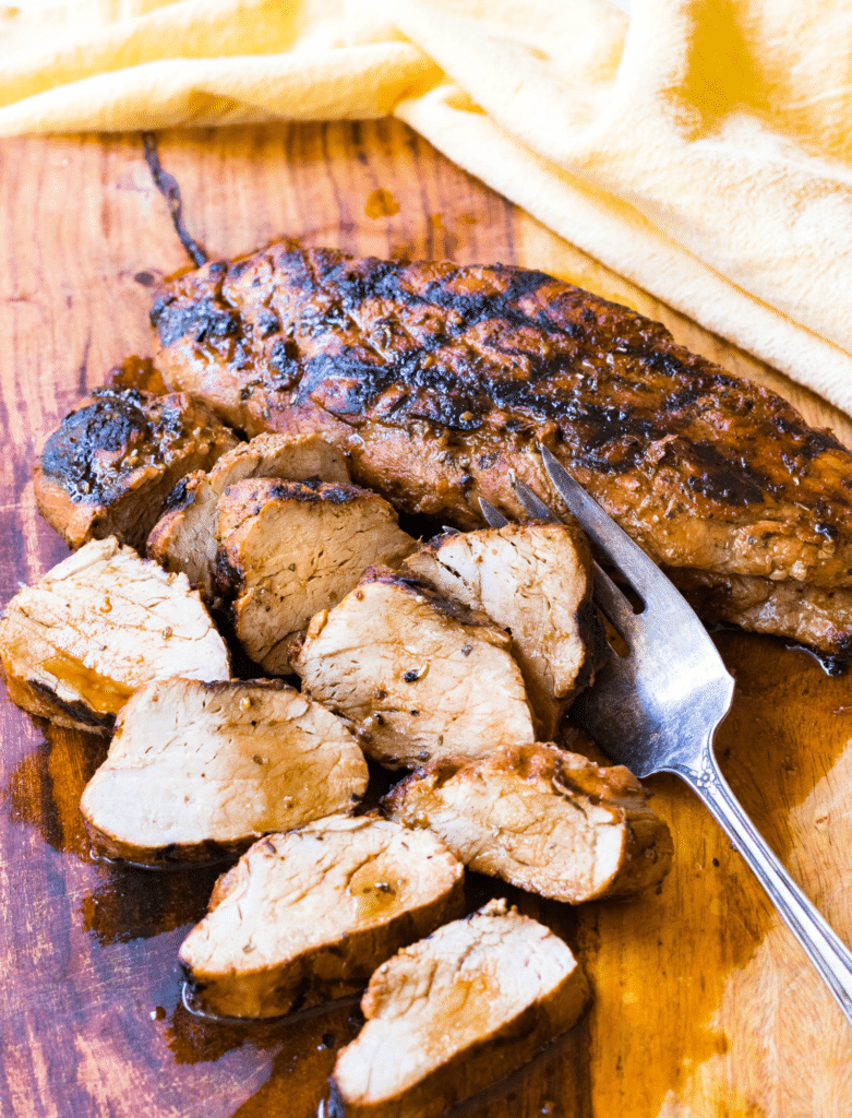 Juicy, succulent, Grilled Pork Tenderloin marinated in olive oil, balsamic vinegar, dried Italian seasoning and garlic, grilled to perfection, sliced, and sitting on a cutting board.