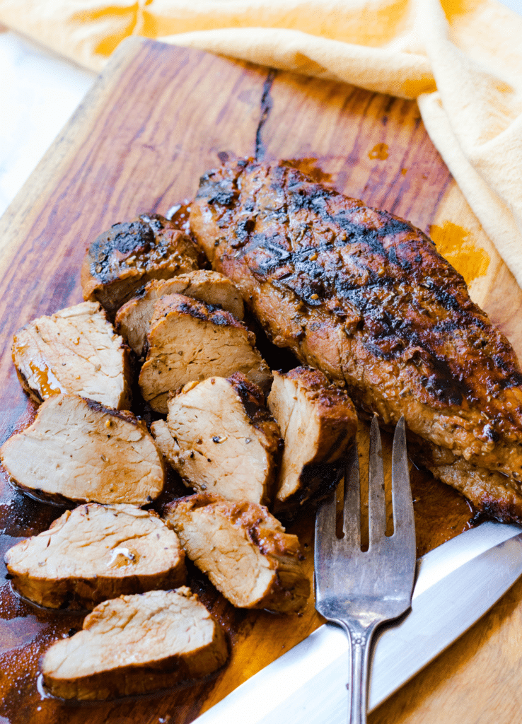 Juicy, succulent, Grilled Pork Tenderloin marinated in olive oil, balsamic vinegar, dried Italian seasoning and garlic, grilled to perfection, sliced, and sitting on a cutting board.