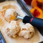 Peach ice cream 3 scoops in black container with scoop and fresh peaches nearby on counter