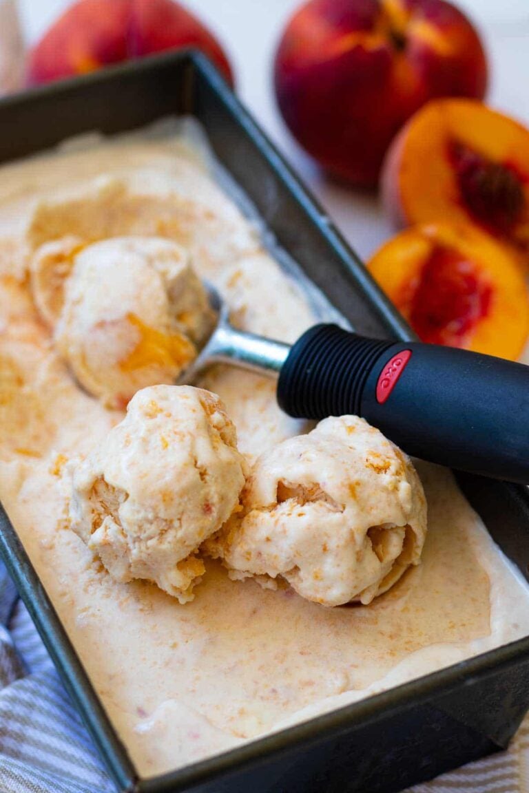 Peach ice cream 3 scoops in black container with scoop and fresh peaches nearby on counter