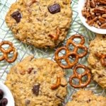 Kitchen Sink Cookies cooling on wire racks with chocolate chips, pecans, and pretzel pieces visible on the cookies and in white bowls