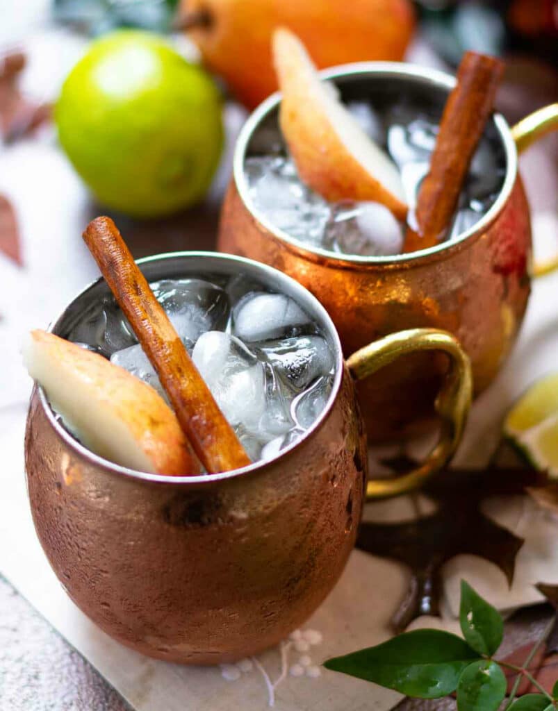 Ice, sliced pears and cinnamon sticks in copper mugs filled with Spiced Pear Moscow Mule drink.